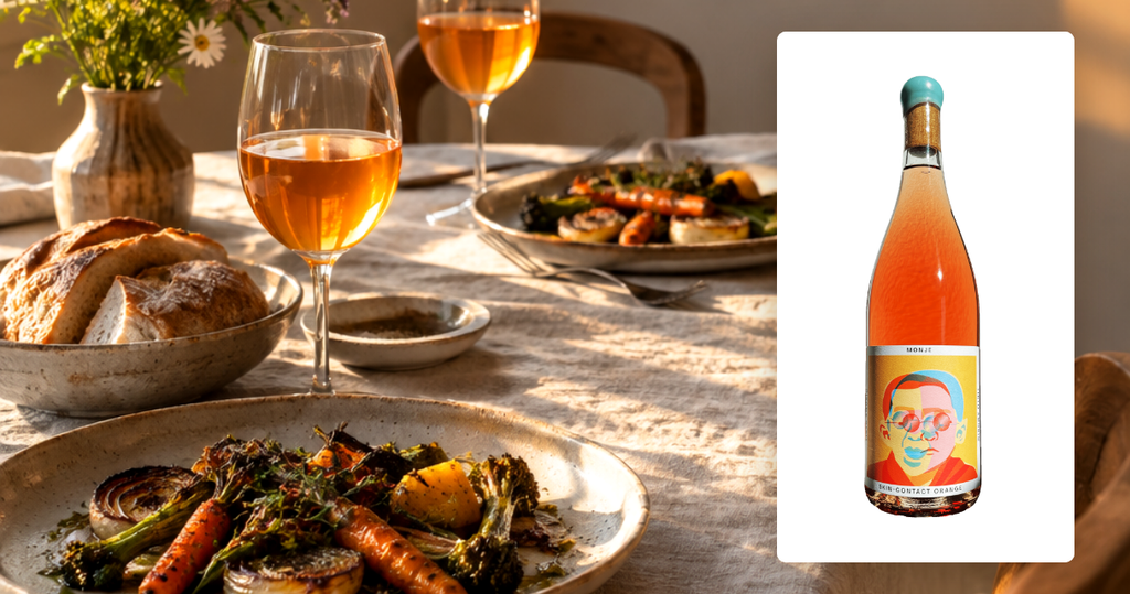 Monje orange wine bottle on a white panel beside a golden-hour dinner table with roasted vegetables and glasses of amber orange wine